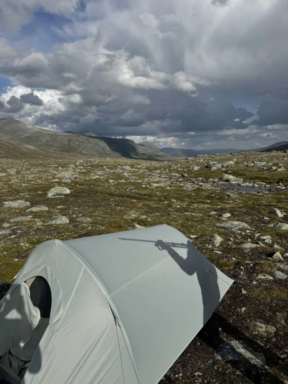Tent camp amid rocky terrain under cloudy skies near a faint rainbow